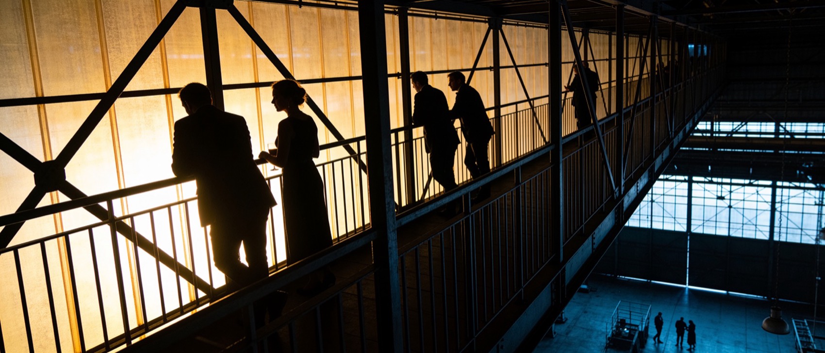 De Gaanderij: gasten als silhouetten tegen warm geel licht op de stalen loopbrug rond de hal, rechts onderin de blauw verlichte halvloer met kleine figuren.