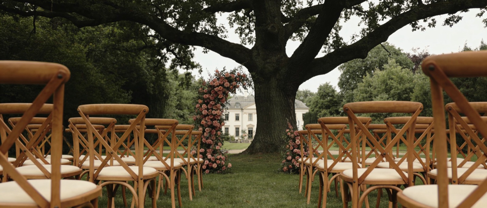 Buitenceremonie gezien vanaf de achterste rij: houten stoelen in het gras, een bloemenboog met rozen onder een oude boom en het landhuis daarachter.