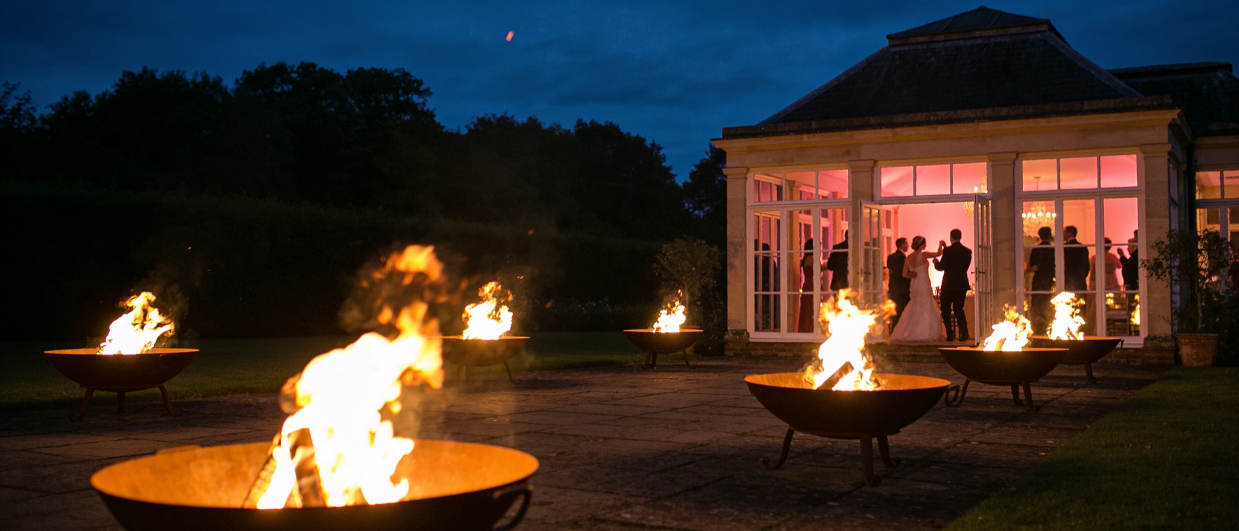 Het voorplein bij avond: brandende vuurkorven op de terrastegels, rechts de verlichte tuinzaal met dansende gasten achter openstaande deuren, daarboven een donkerblauwe lucht.