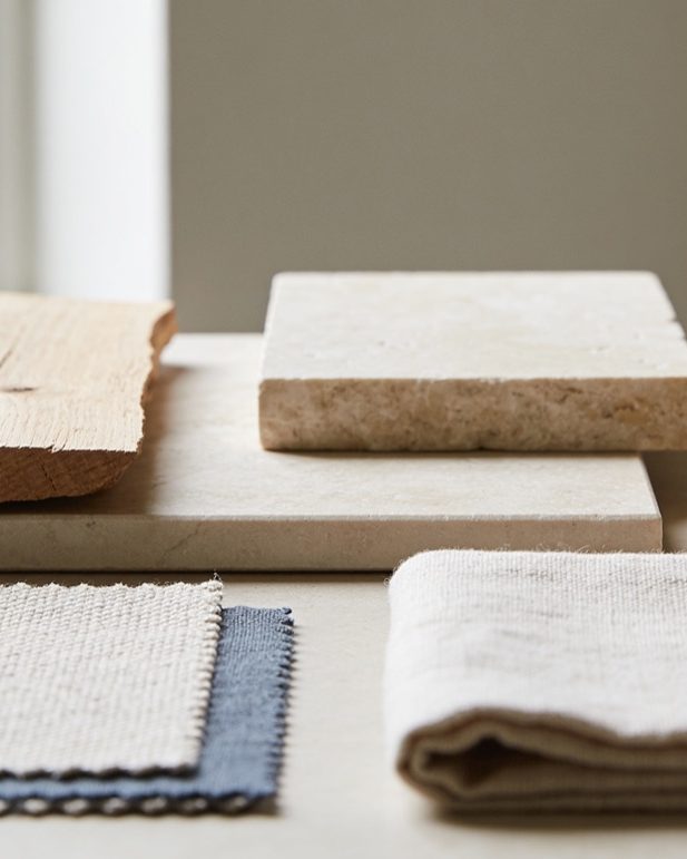 Materiaalstalen op een werktafel bij het raam: een ruw stuk eiken, twee tegels kalksteen en gevouwen linnen en wollen lappen.