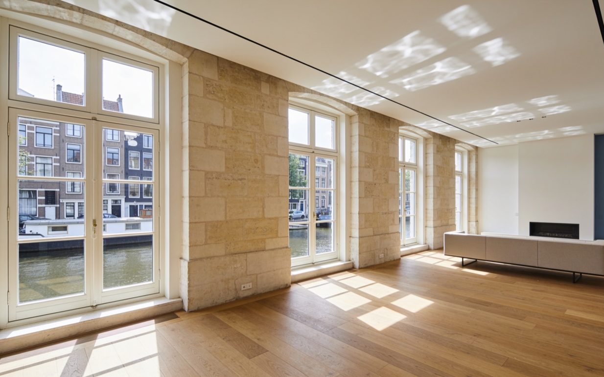 Lege woonkamer in een grachtenpand met hoge stenen kozijnen, uitzicht op het water en een eiken vloer met lichtvlekken.
